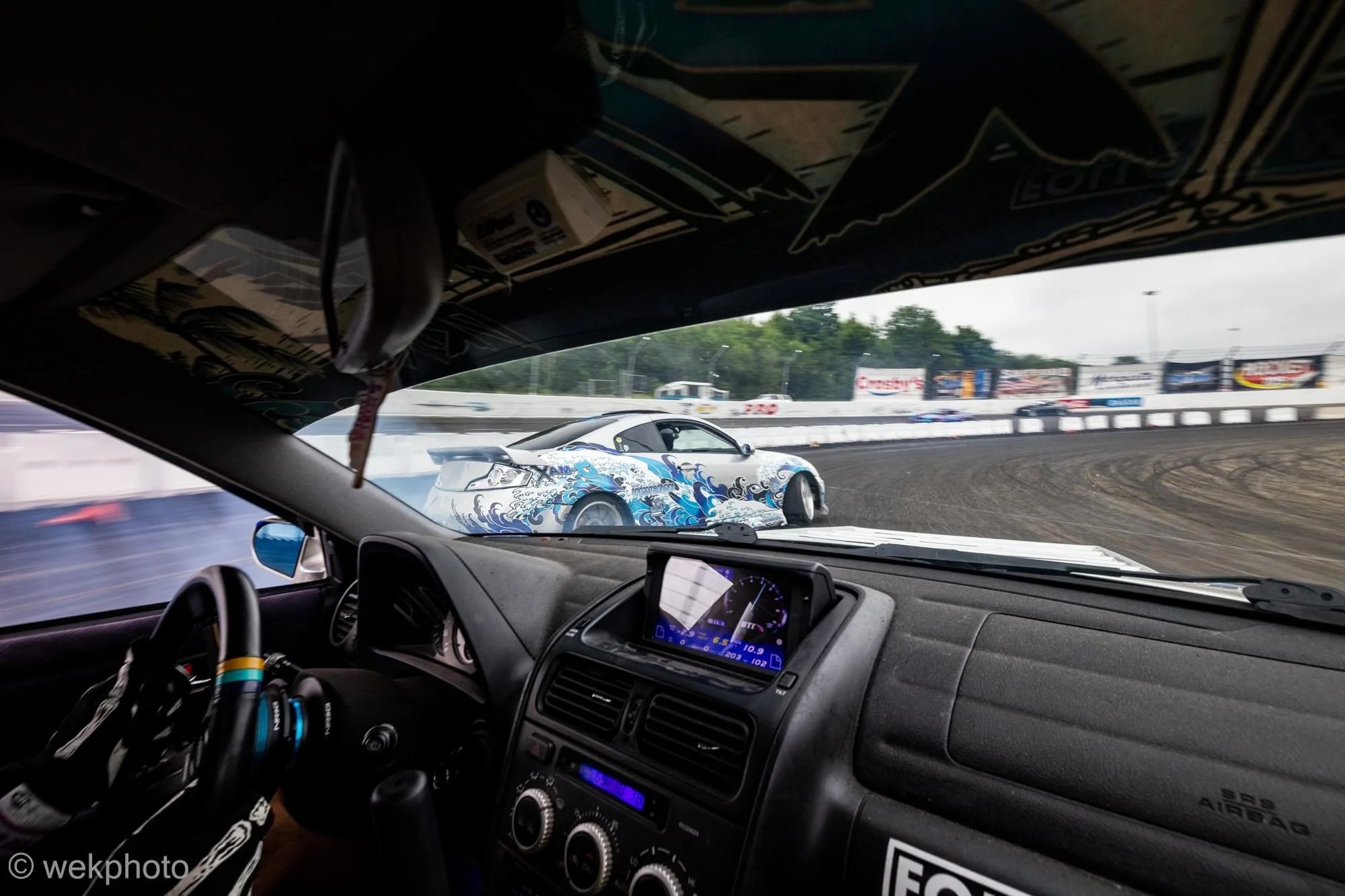 Inside a race car, looking out at a blue and white race car on a dirt track, with a digital dashboard display in the foreground and advertisements on the fence in the background.