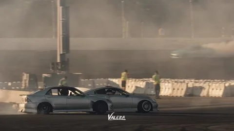 A silver sports car drifting on a racetrack with smoke and dust in the air, and three safety personnel nearby.
