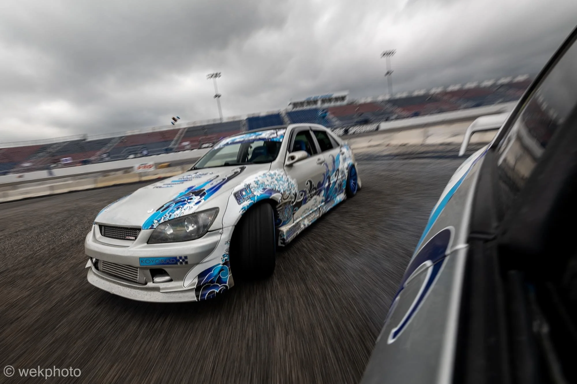 A white race car with blue and black graphics drifting on a race track under cloudy skies, with stadium seating in the background.