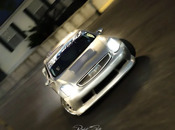 Silver sports car on a dark street at night, with a building and plants in the background.