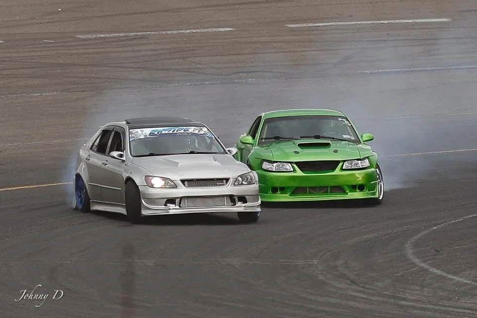 Two race cars, one silver and one green, drifting on a race track with smoke coming from their tires.