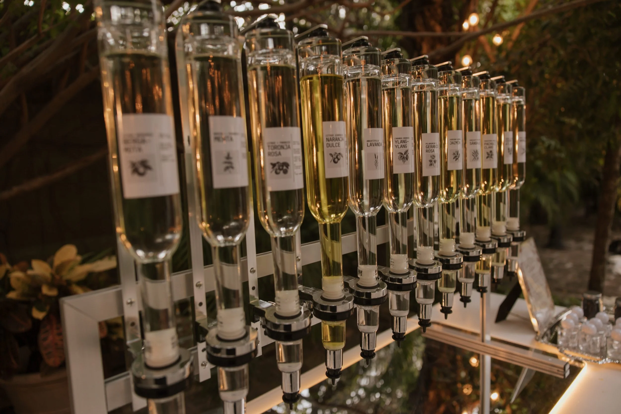 A fountain display of vertical glass vials filled with various liquids, with labels indicating different scents, arranged on a white stand at an outdoor event during the evening.