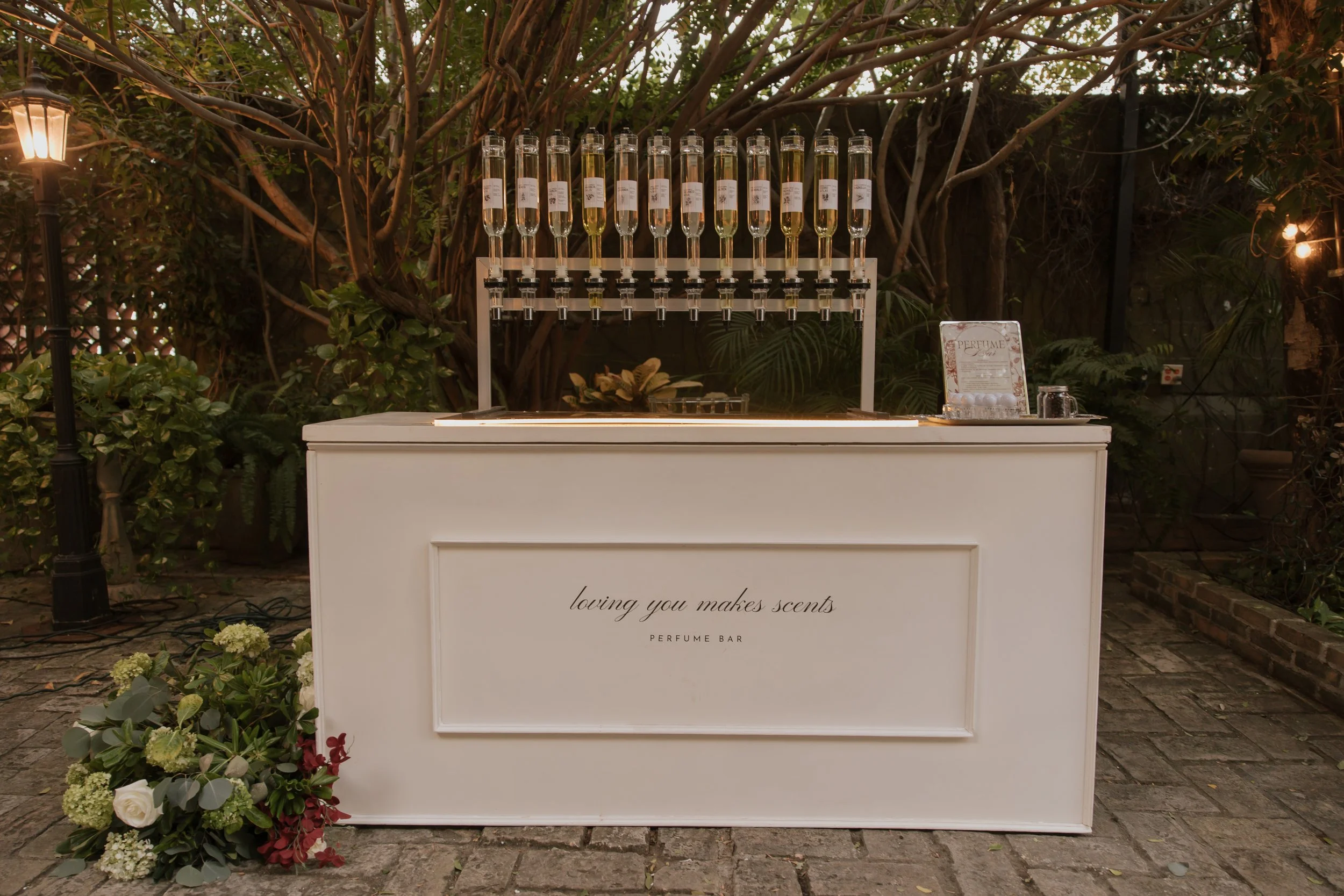 An outdoor perfume bar setup with a white counter reading 'loving you makes scents PERFUME BAR' in front of a garden with trees and shrubs. The counter displays bottles of perfume and a sign with perfume information. A floral arrangement is positioned on the ground to the left of the counter.