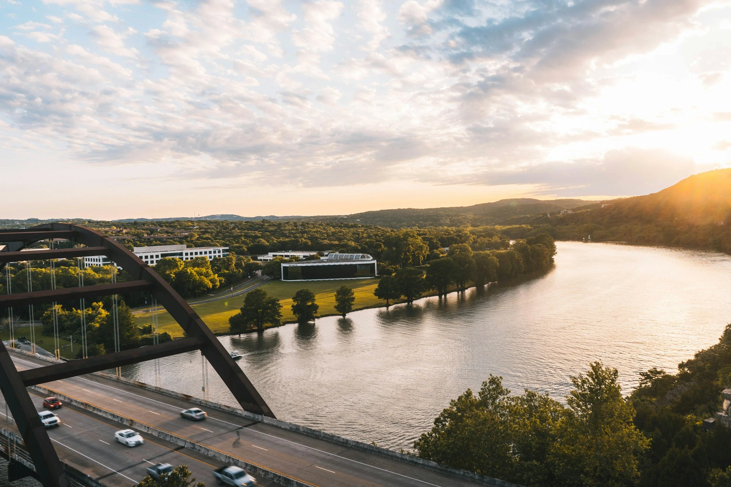 View of a river flowing through a green landscape with trees, a bridge with cars, and buildings in the distance during sunset.