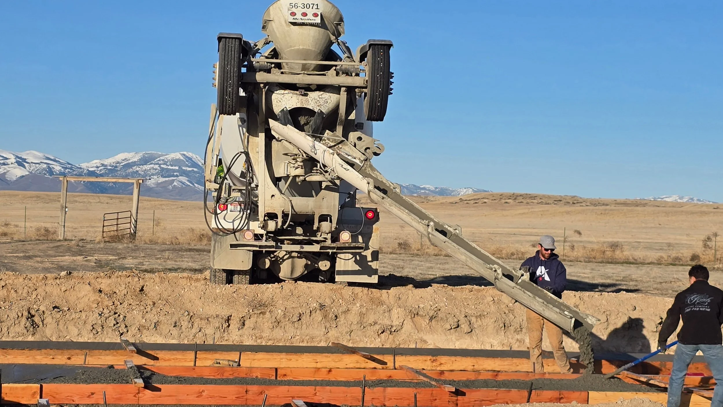 Two construction workers are pouring concrete from a cement mixer truck into wooden forms on a construction site, with a rural landscape and snow-capped mountains in the background.