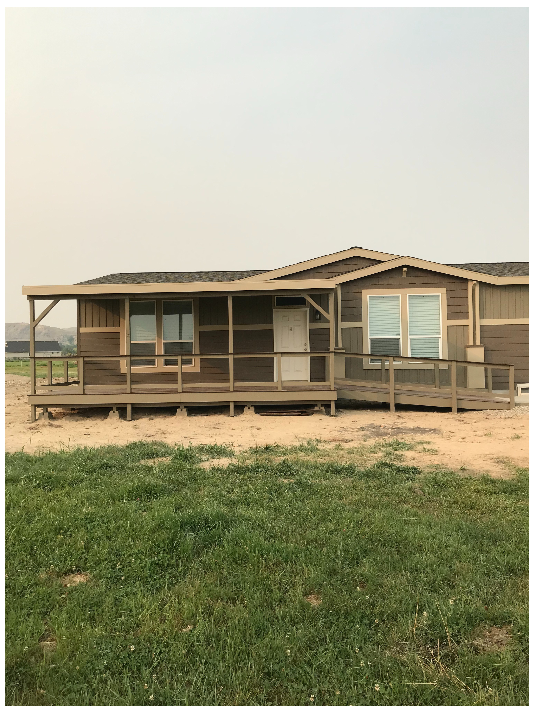 A small, single-story house with a front porch, brown siding, and two large windows, situated on a grassy yard with some patches of dirt.