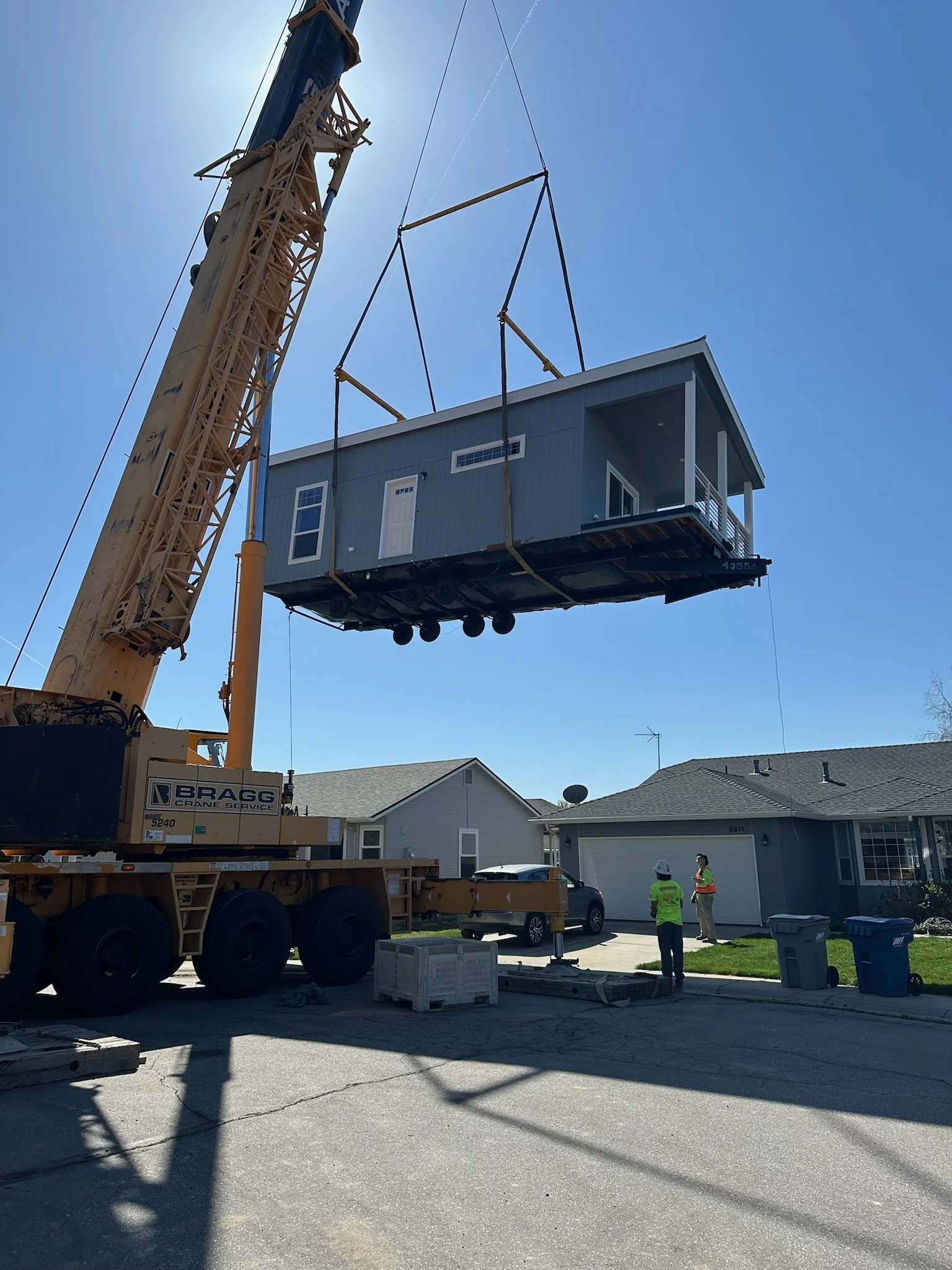 A house is being lifted by a crane into a neighborhood with single-story homes. Two workers in safety vests are standing nearby, overseeing the operation on a sunny day.