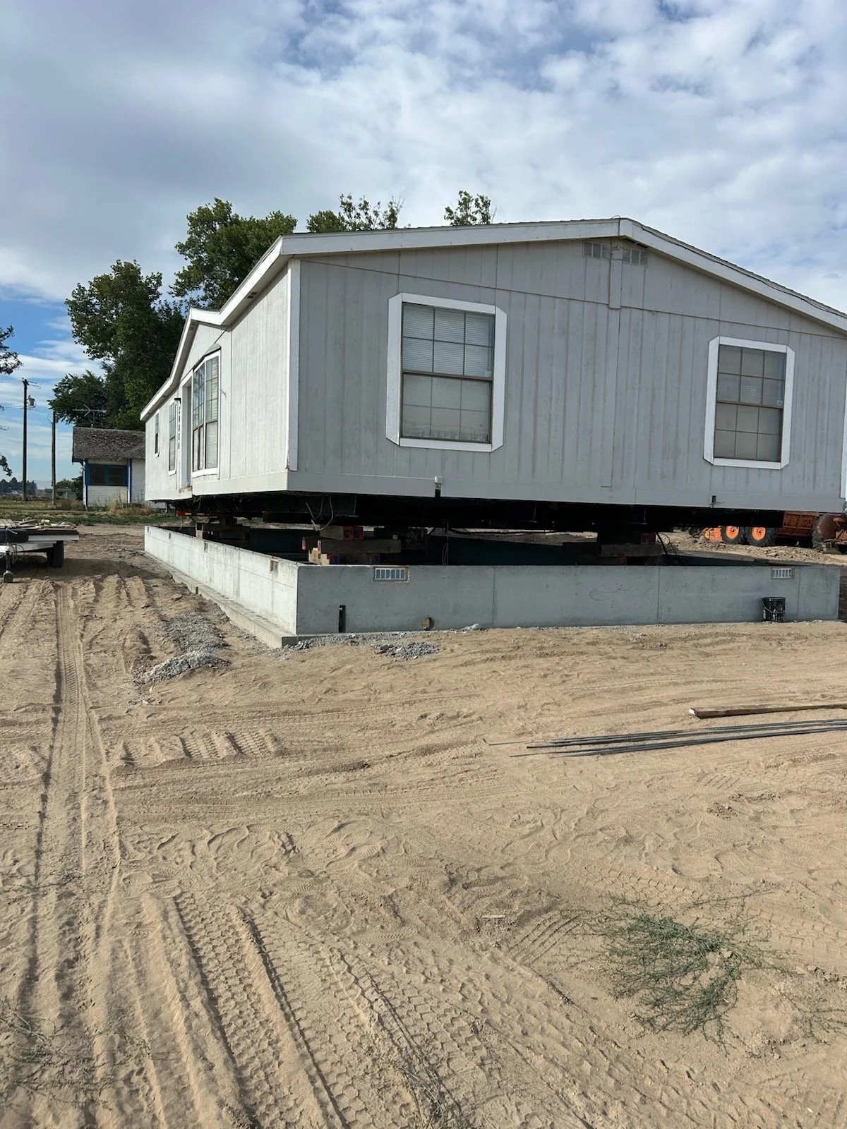 A house is elevated on a foundation on a construction site with tire tracks in the dirt and blue sky with clouds overhead.