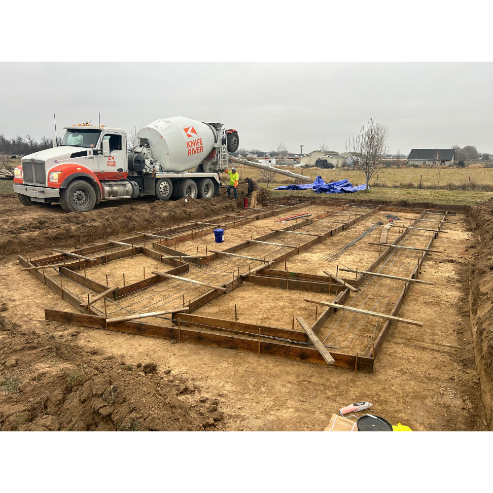 Construction site with wooden framing and rebar for a building foundation, a cement mixer truck labeled 'Knife River', and workers installing the foundation on an overcast day in a rural area.