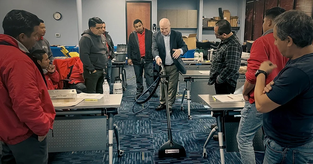 A man named Jeff Cutshell demonstrating a cleaning or inspection device to a group of people in an office or classroom setting, with tables, papers, and equipment around.