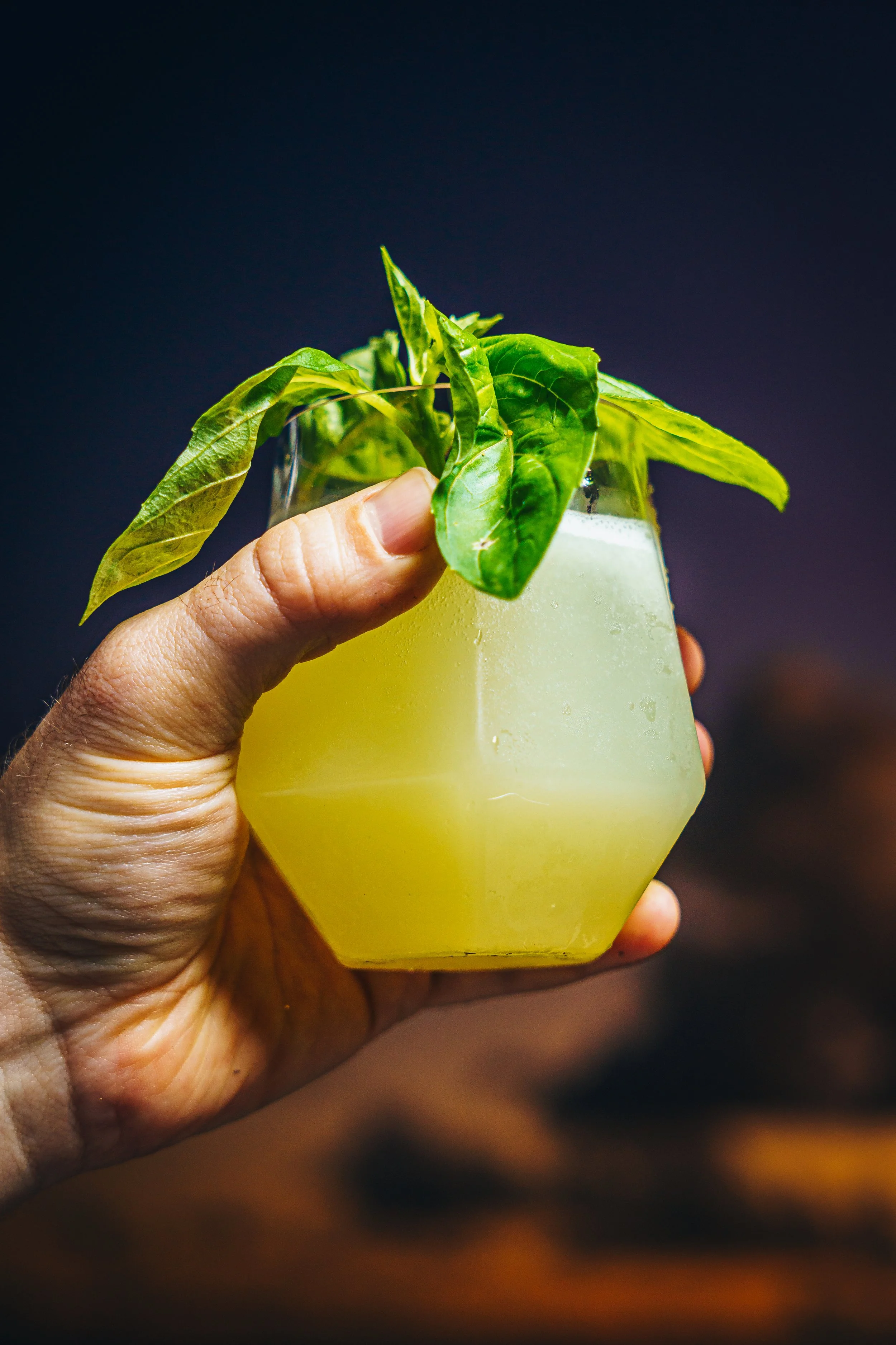 Close-up of a hand holding a glass of yellow-green beverage topped with fresh green herbs against a dark background.