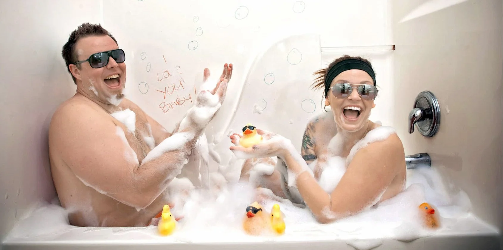 A man and woman wearing sunglasses and enjoying a bubble bath with rubber ducks, smiling and having fun.