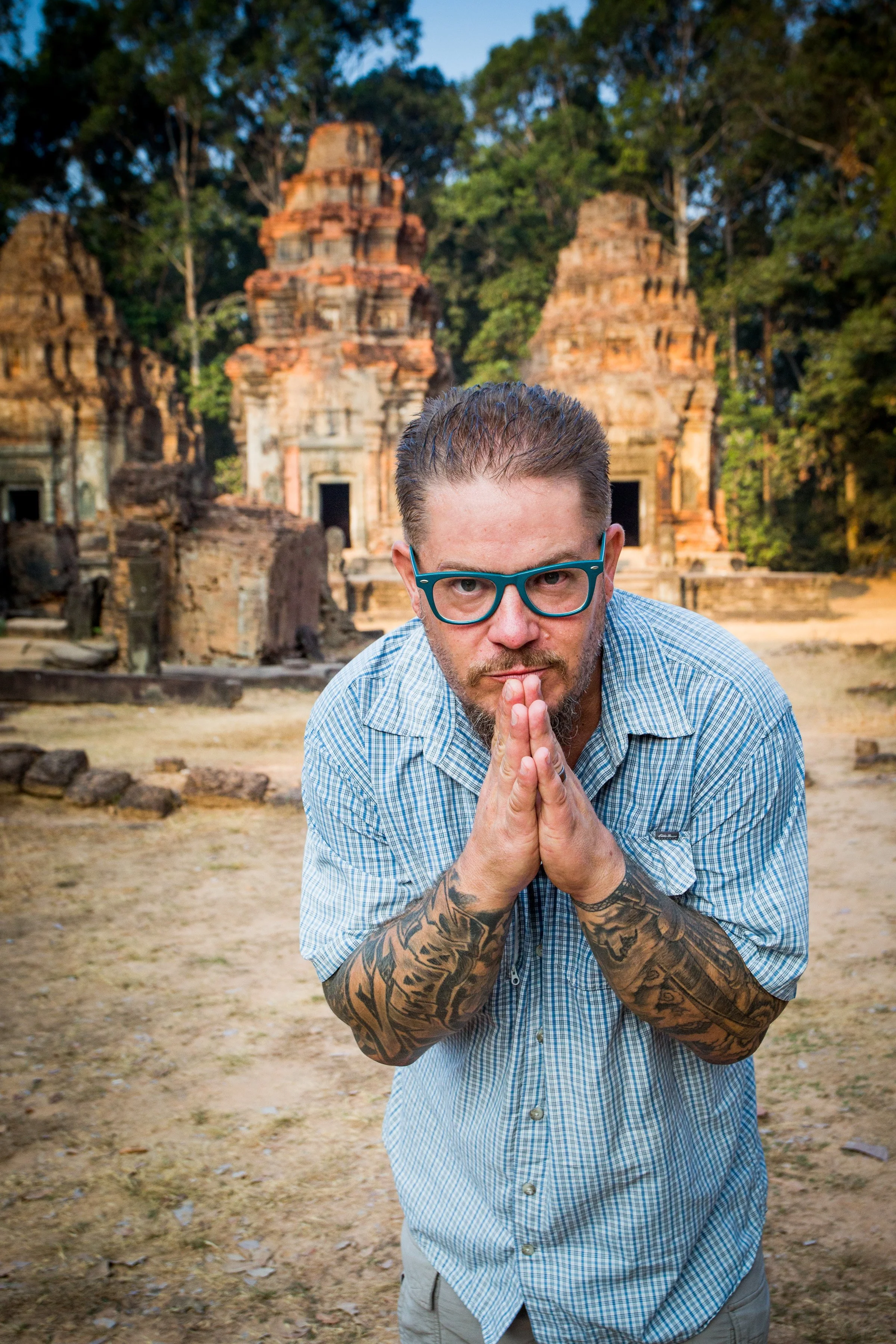A man with tattoos on his arms, blue glasses, and short brown hair, dressed in a light blue checkered shirt, is kneeling with hands pressed together in a prayer-like gesture in front of ancient ruins with a forest background.