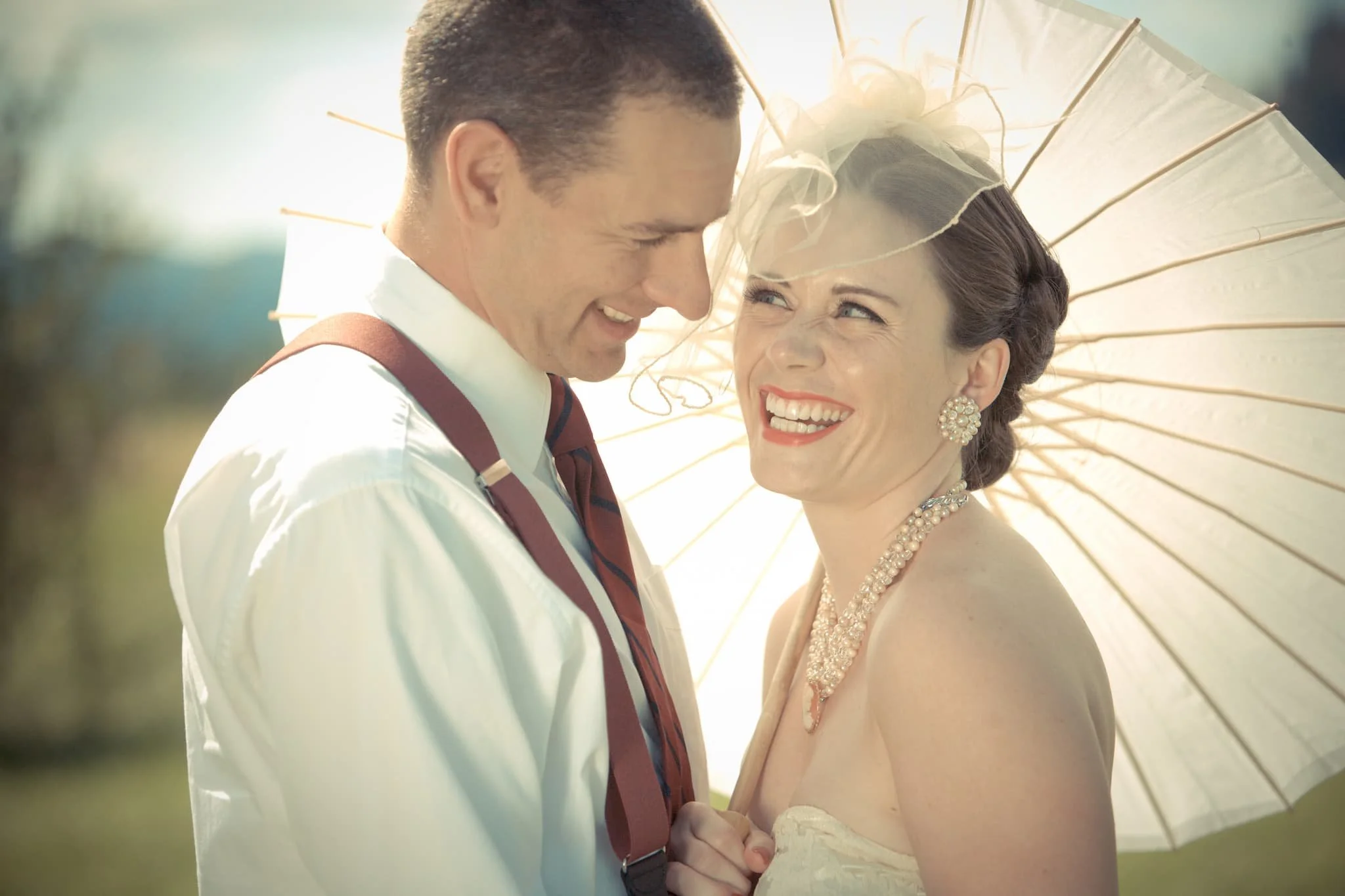 A smiling bride and groom standing close beneath a white parasol outdoors, with sunlight illuminating their faces.
