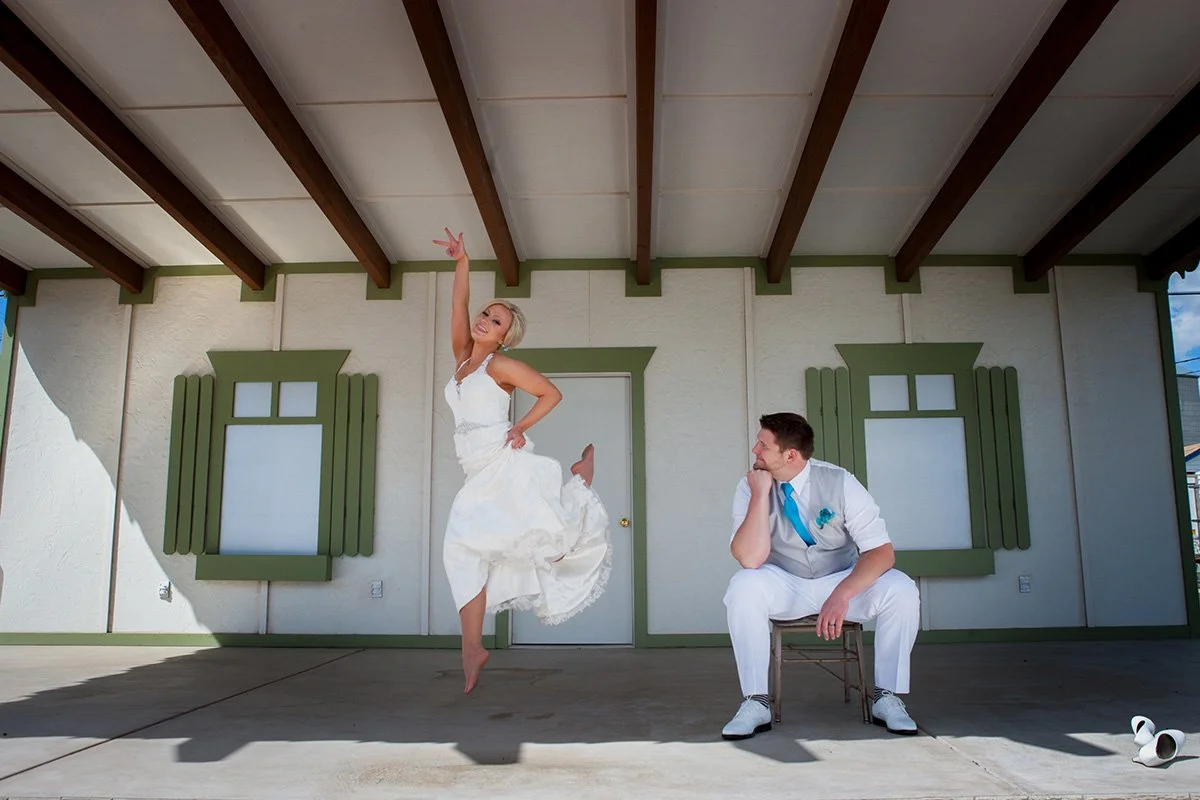 A woman in a white dress jumping joyfully while a man in a white suit with a blue tie seated on a stool looks on, set against a building with green window frames and a door, under a wooden-beamed ceiling.