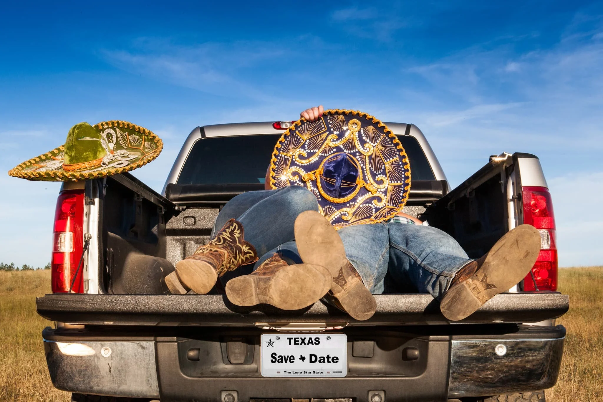 People lying in the back of a pickup truck with sombreros, cowboy boots, and jeans, on a grassy field under a blue sky.