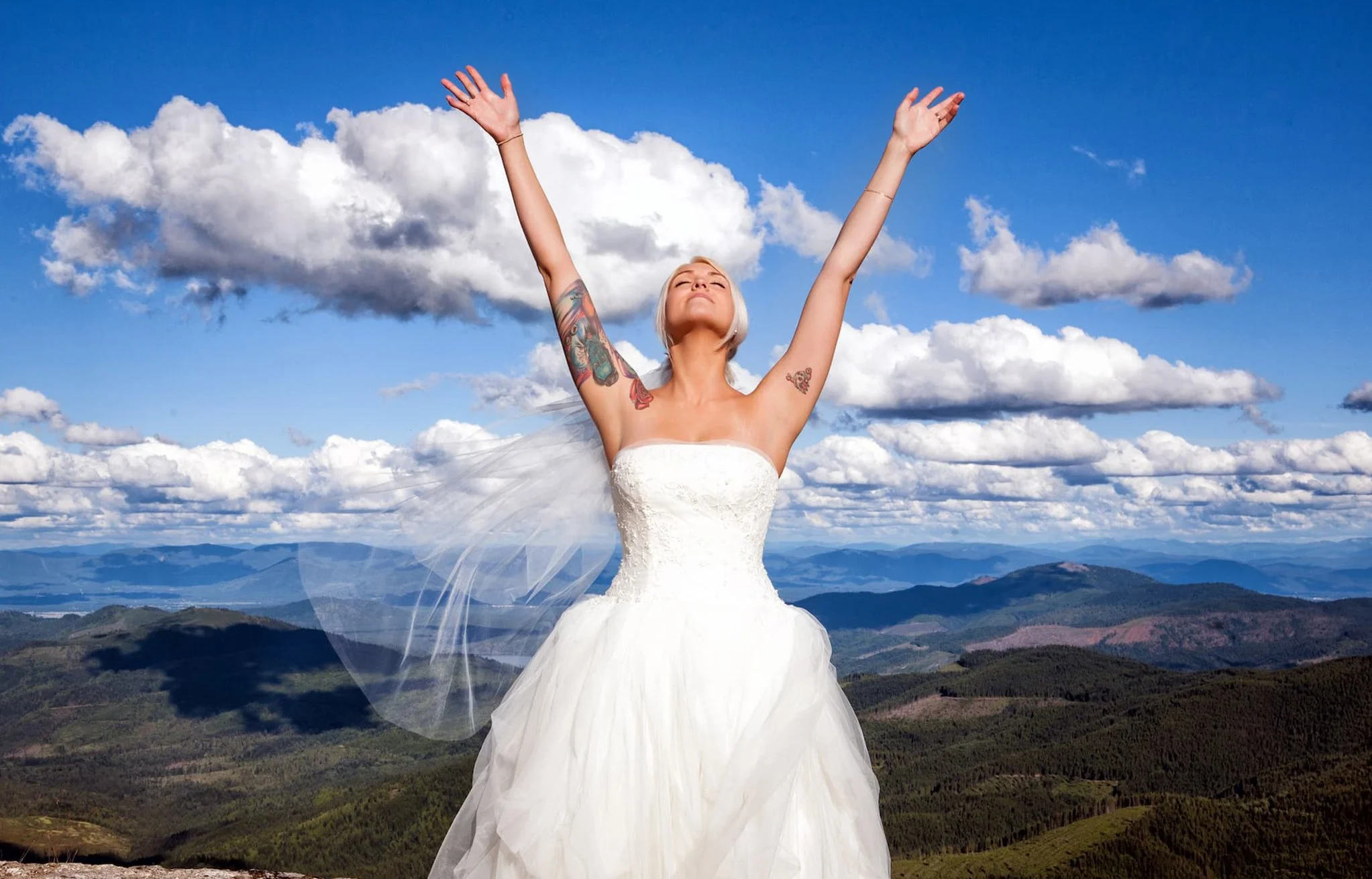 A woman in a wedding dress with tattoos on her arms, standing on a mountain with arms raised and eyes closed, under a blue sky with clouds.