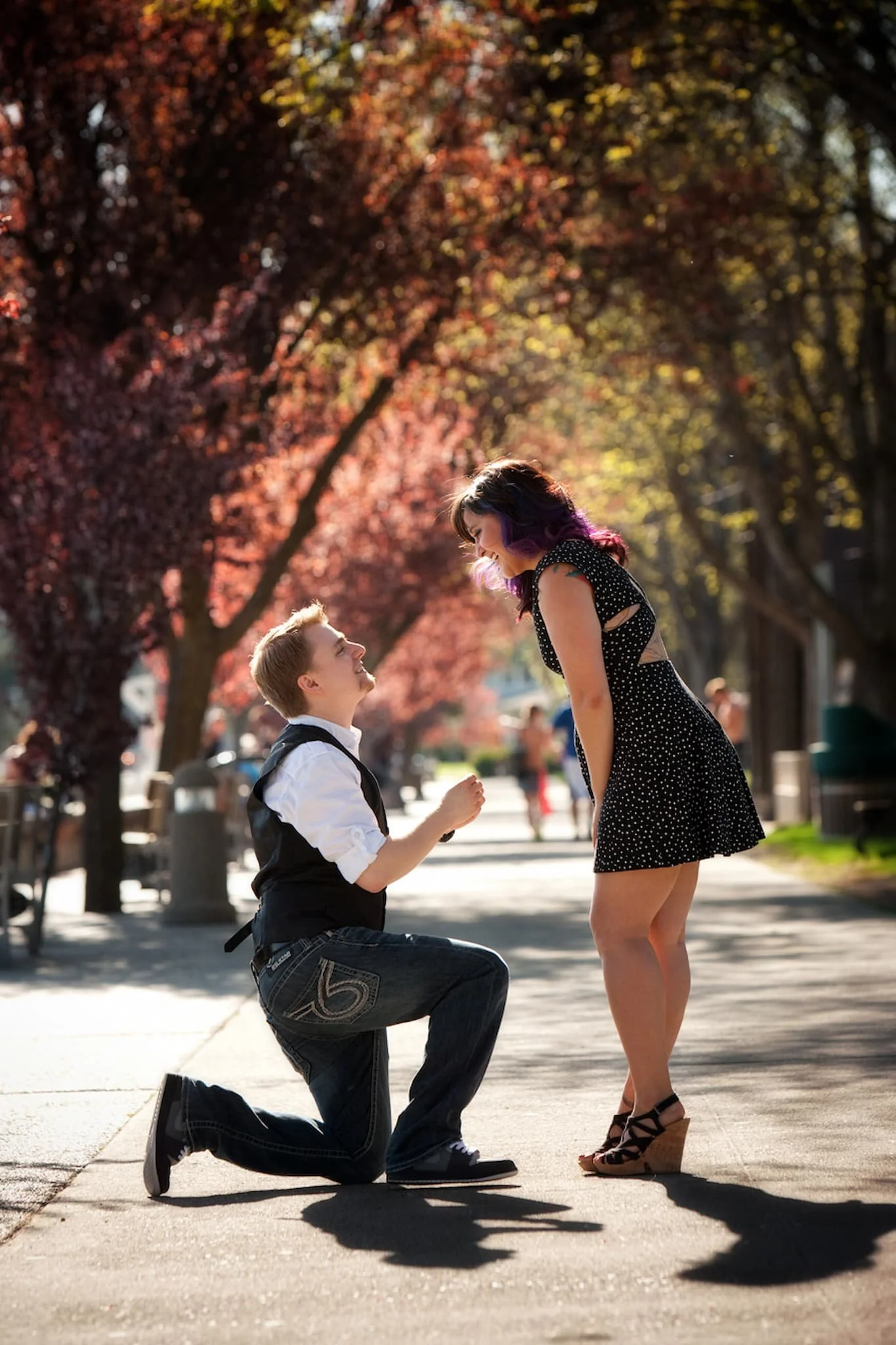 A man proposing marriage to a woman on a park sidewalk with trees and autumn foliage in the background.
