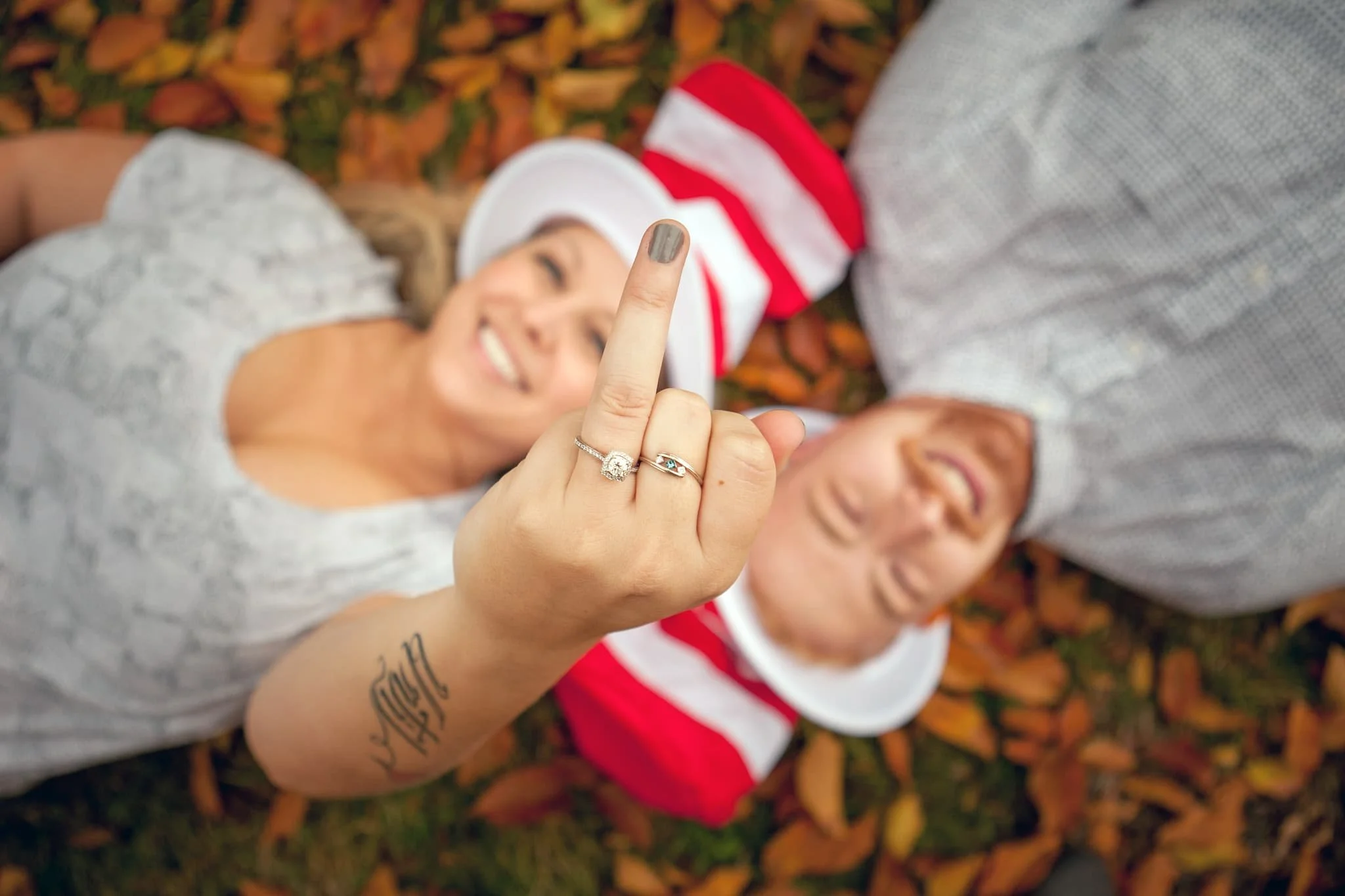 A woman lying on the ground with autumn leaves, showing her middle finger with a wedding ring and a tattoo on her forearm, smiling and a man next to her.