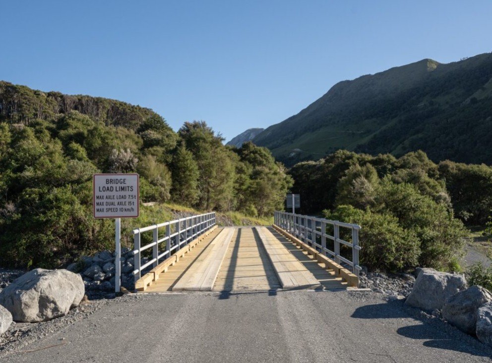 A small bridge with safety railings over a creek, surrounded by green hills, with a bright blue sky. There is a sign on the left indicating load limits and speed restrictions.