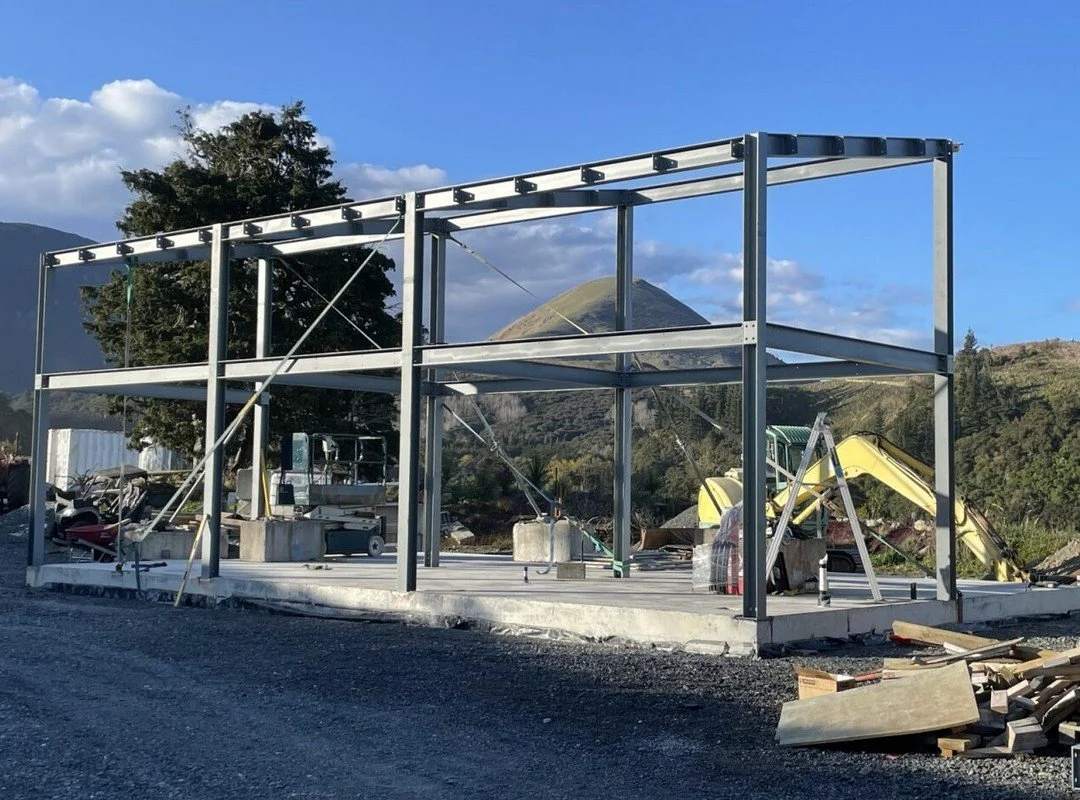 Construction site with a steel frame structure being assembled on a concrete foundation, with construction equipment and materials around, mountainous landscape in the background.