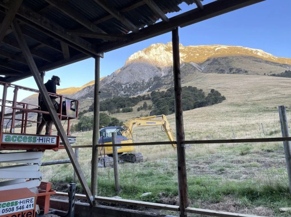 A person operating a small hydraulic excavator on a grassy field with a mountain in the background, viewed from inside a wooden structure with scaffolding labeled 'ACCESS-HIRE'.