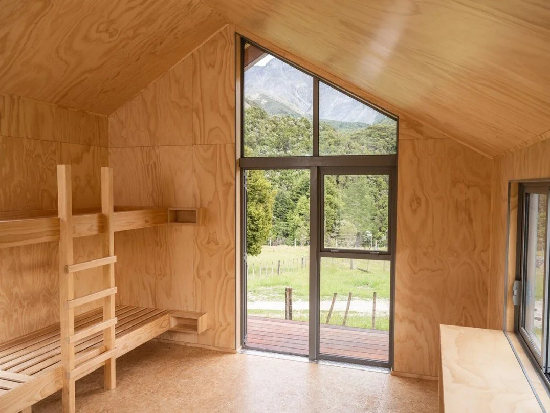 A small room with wooden walls and sloped ceiling, featuring a bunks bed on the left, a large window with a view of trees and mountains, and a small wooden bench under the window.