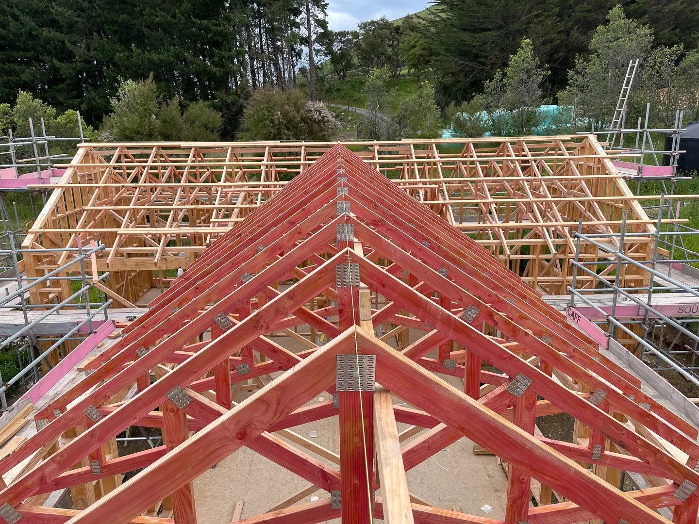 Construction site with a wooden roof frame being built, surrounded by scaffolding, overlooking a green outdoor area with trees and hills.