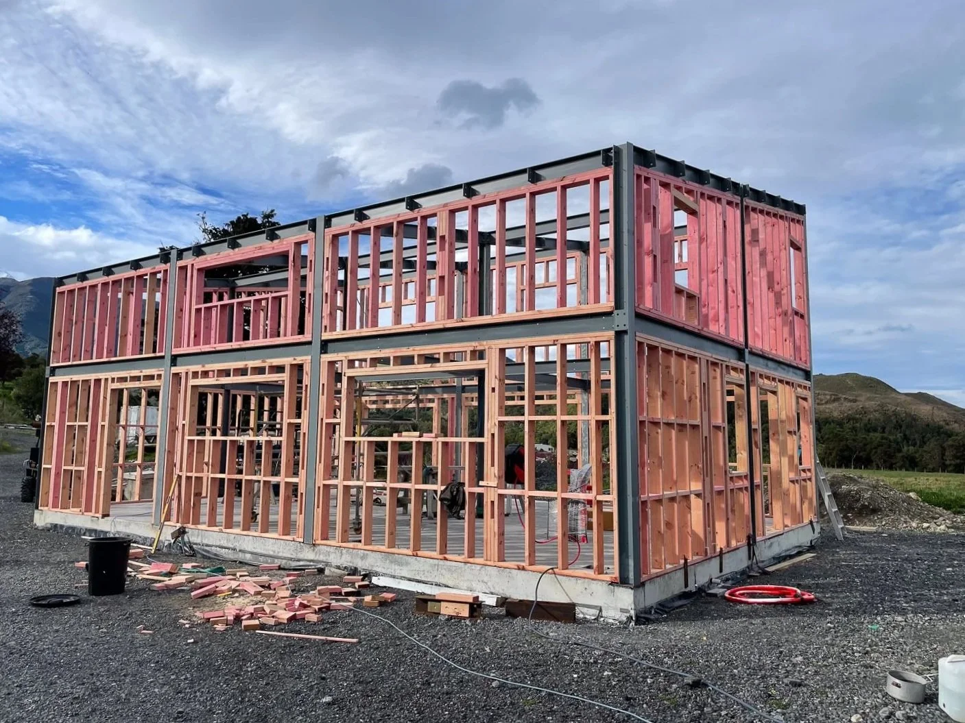 A two-story house under construction with wooden framing, set on a gravel foundation, with mountains and a cloudy sky in the background.