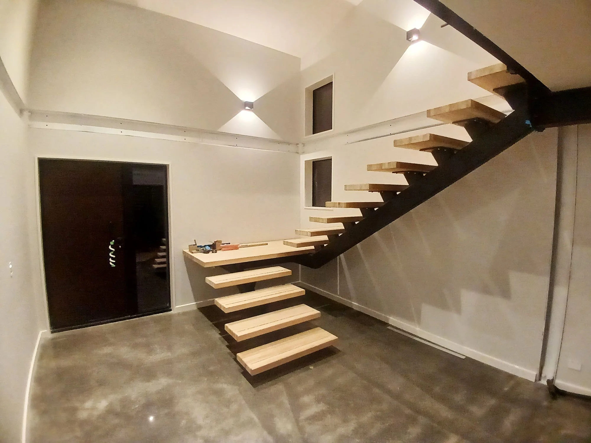 Interior view of a modern staircase with floating wooden steps and a black support structure, set against white walls with minimal lighting.
