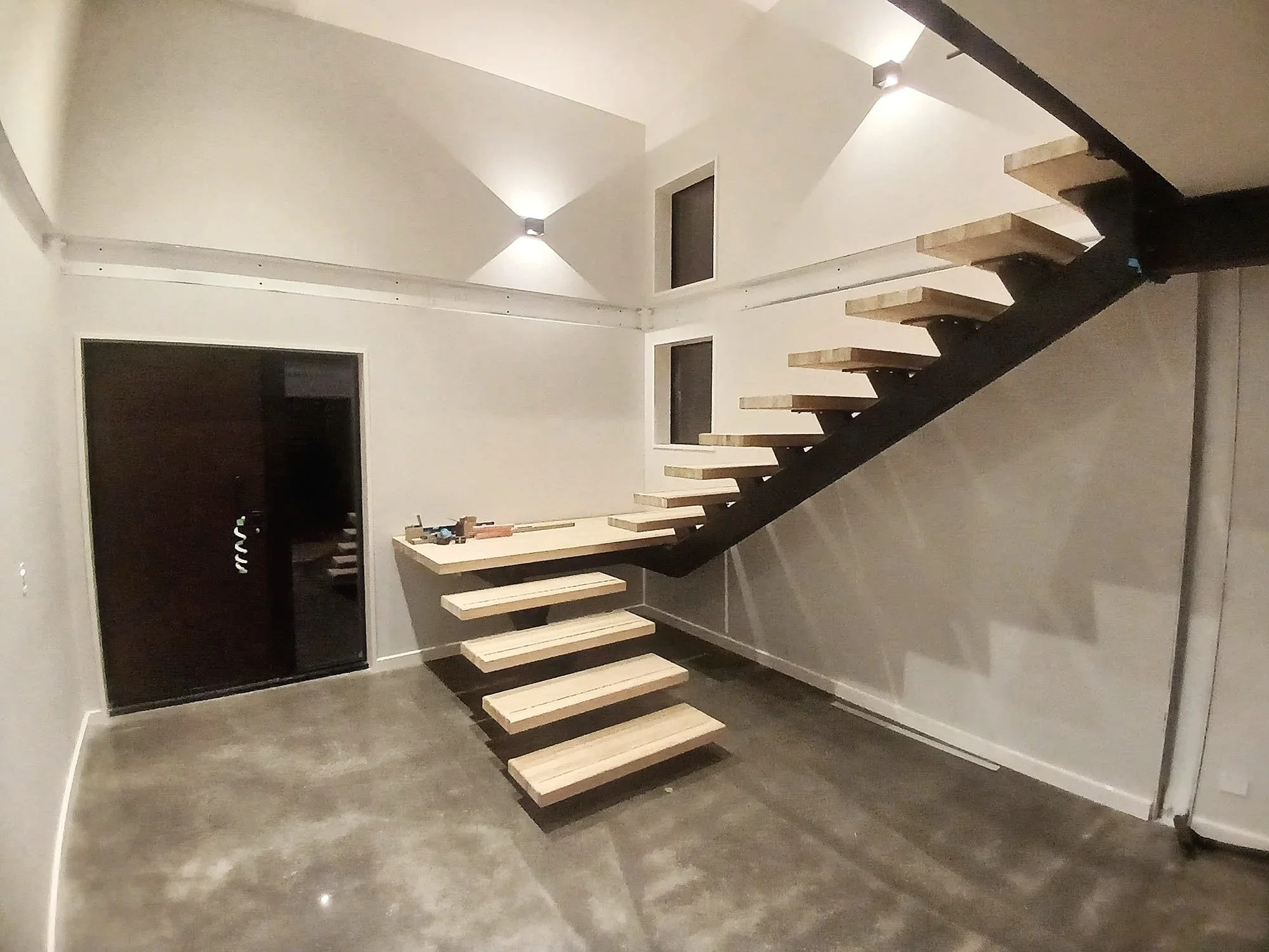 Interior of a modern room with a black floating staircase made of wood steps and a black steel support, leading to an upper level. The room has white walls, with two small windows and a door. The floor appears to be made of dark polished concrete or similar material, and the ceiling has spotlights.
