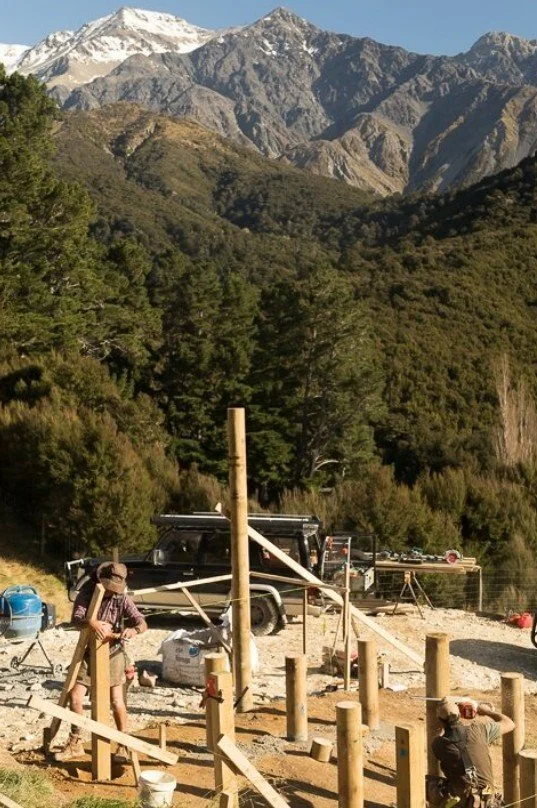 People working on constructing wooden posts and beams outdoors, with forested mountains and snow-capped peaks in the background.
