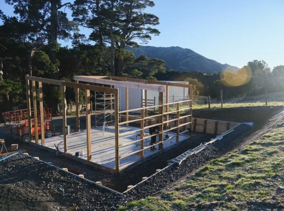 Construction site with wooden framing, scaffolding, and building materials, set against a backdrop of trees and mountains during daylight.