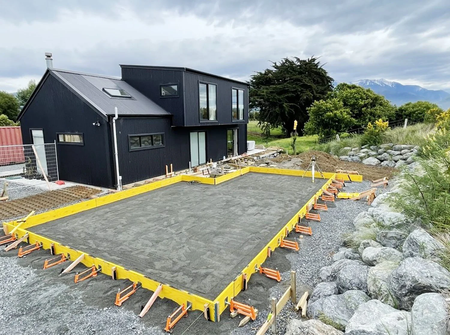A modern black house under construction with concrete foundation framework and construction tools, set in a scenic landscape with trees and mountains in the background.