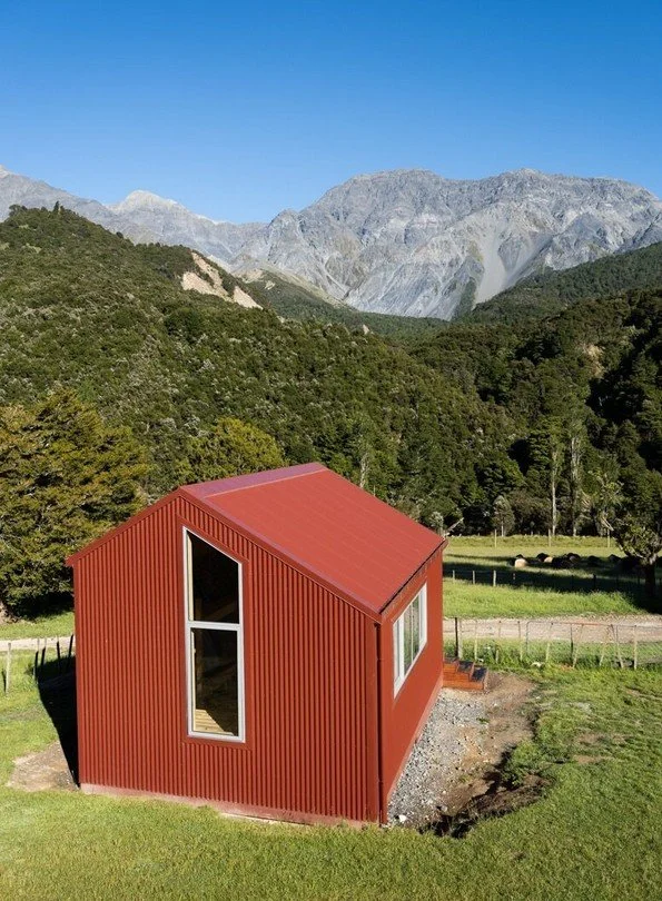 A red modern tiny house with a sloped roof situated on green grass, surrounded by trees and mountains in the background under a clear blue sky.