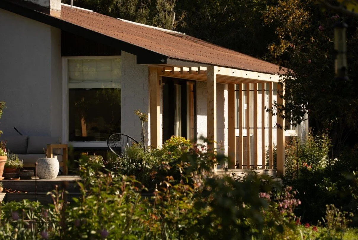 A house with a sloped roof, a large window, and an outdoor seating area surrounded by plants and flowers.