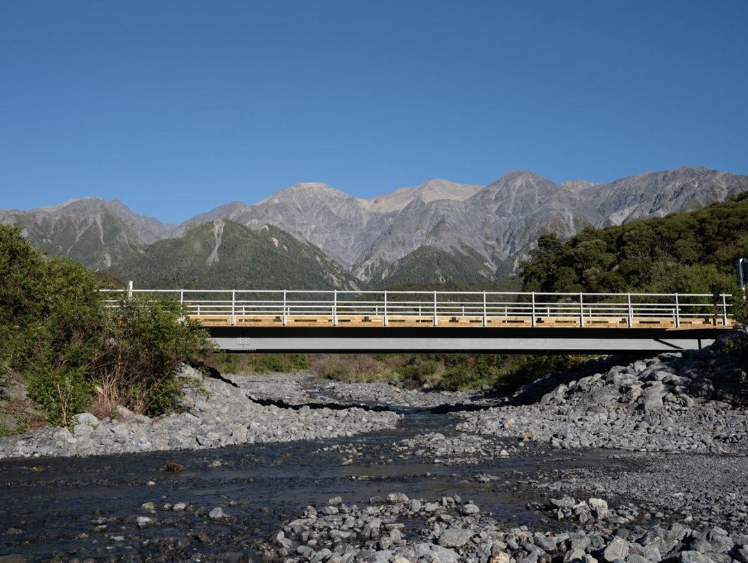 A mountain landscape with a bridge crossing over a rocky riverbed, surrounded by green forests, under a clear blue sky.