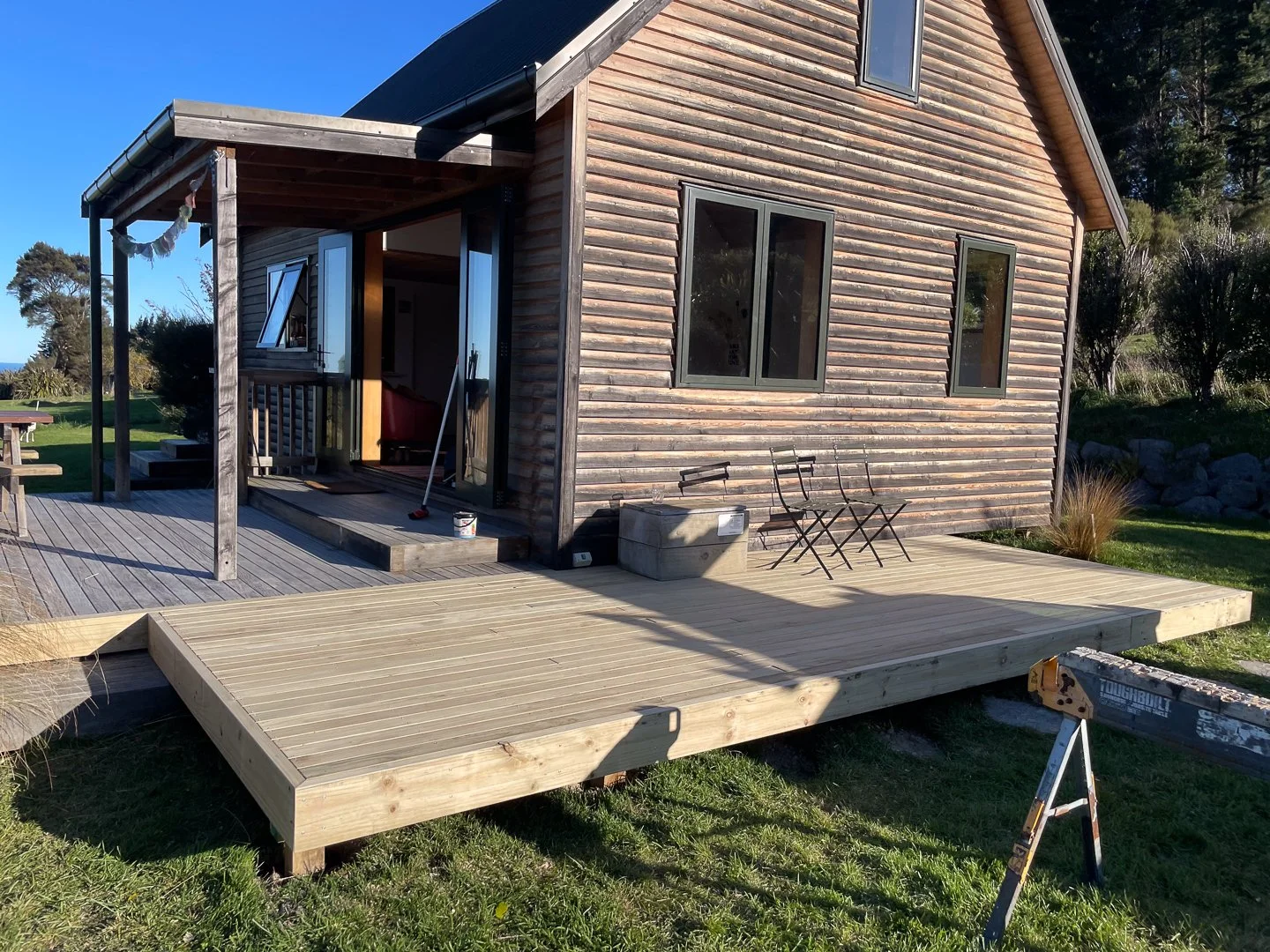 A rural wooden house with a newly constructed wooden deck in front, surrounded by grassy lawn and trees, with construction tools and chairs on the deck.