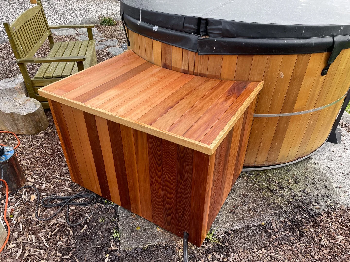 A wooden hot tub with a dark cover and a small wooden storage box nearby, set on a gravel and dirt area with a wooden bench in the background.