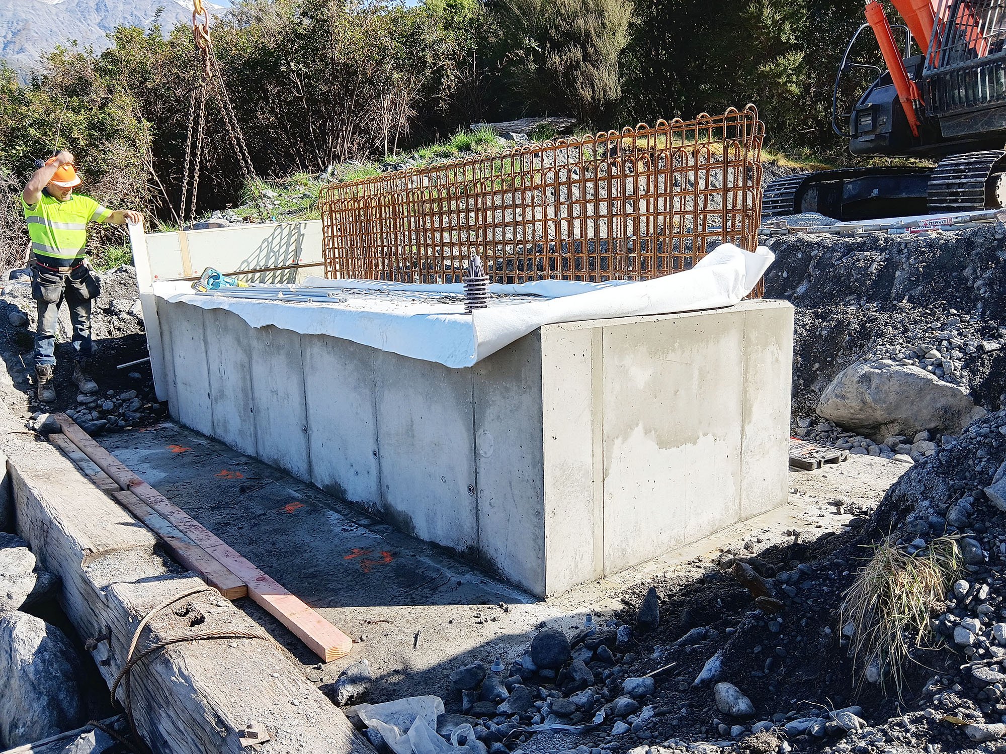 Construction worker in safety gear standing next to a concrete structure with orange safety barriers on top, surrounded by construction tools and equipment at a construction site.