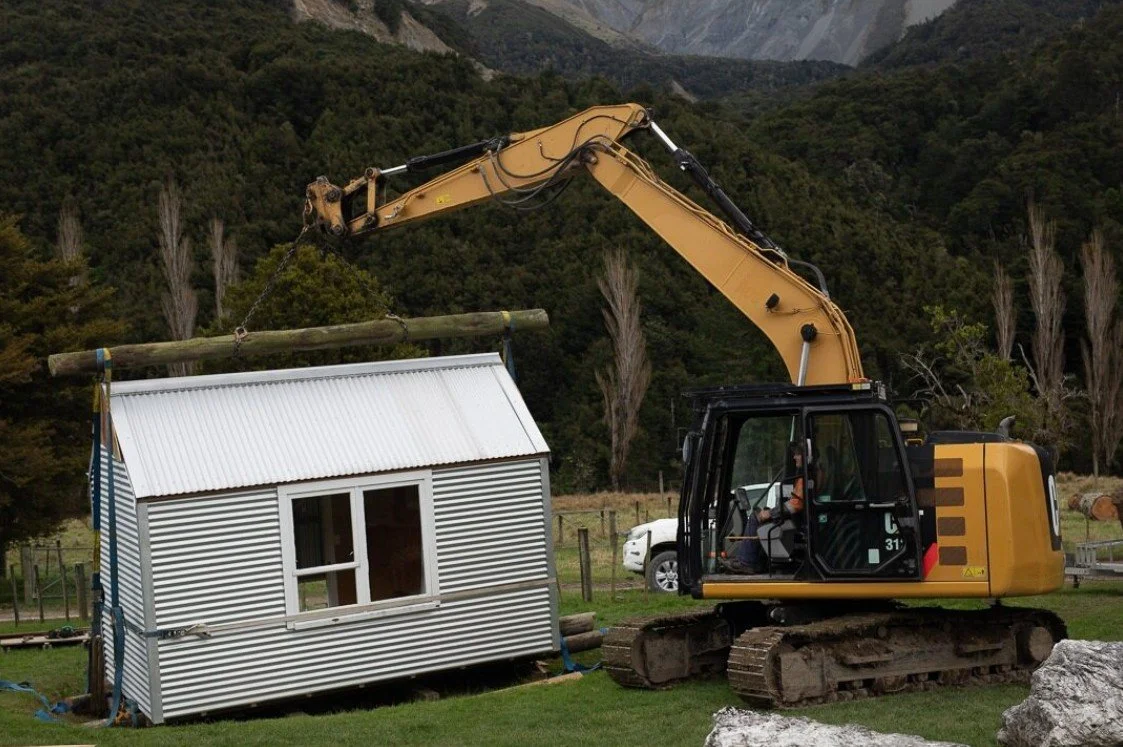 A small construction excavator lifting a wooden log onto a miniature house with metal siding, set in a rural area with mountains and trees in the background.