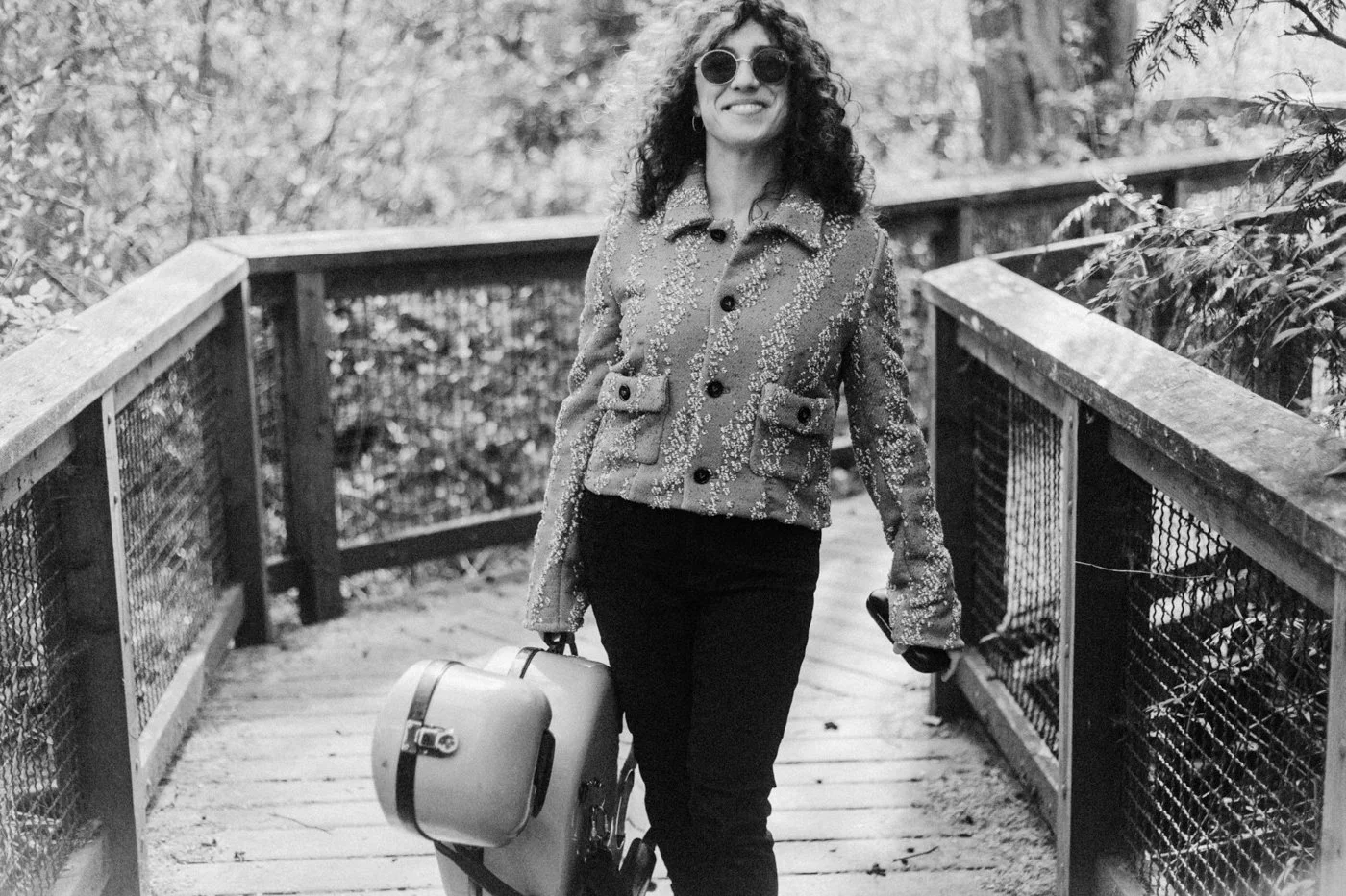 Natalie Mai Hall, a woman with curly hair wearing sunglasses, a patterned jacket, and black pants, walking on a wooden bridge with a cello case in one hand, surrounded by trees.