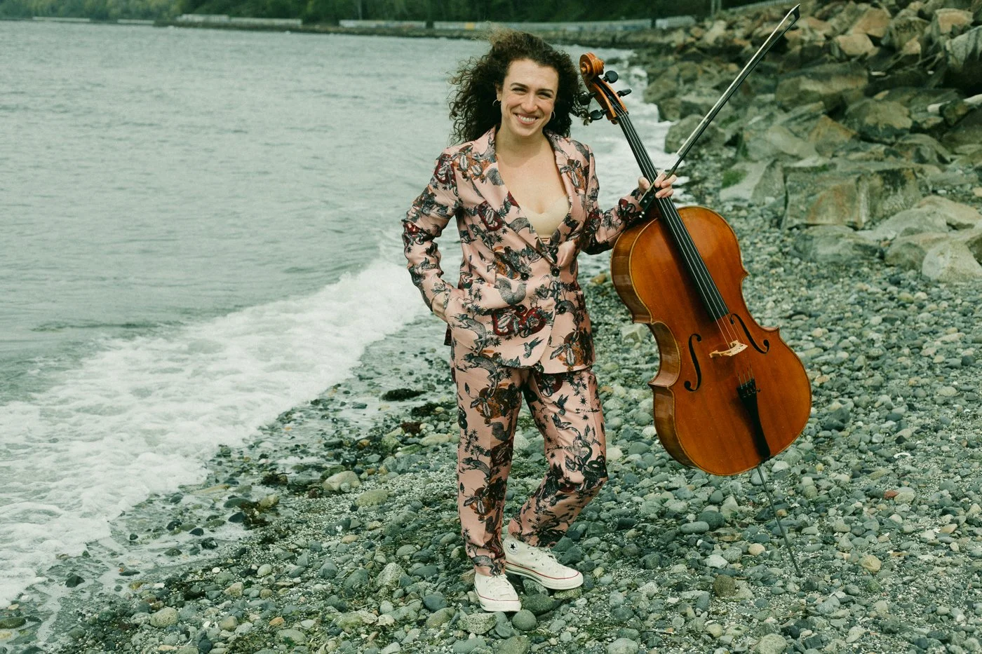 Natalie Mai Hall, a woman with curly hair standing on a pebble beach near the water, holding a cello and smiling at the camera, wearing a matching floral suit and white sneakers.