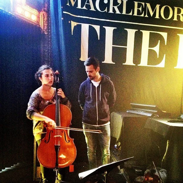 A woman playing a cello and a man standing next to her in a performance venue with stage lights and a large black and yellow sign behind them.