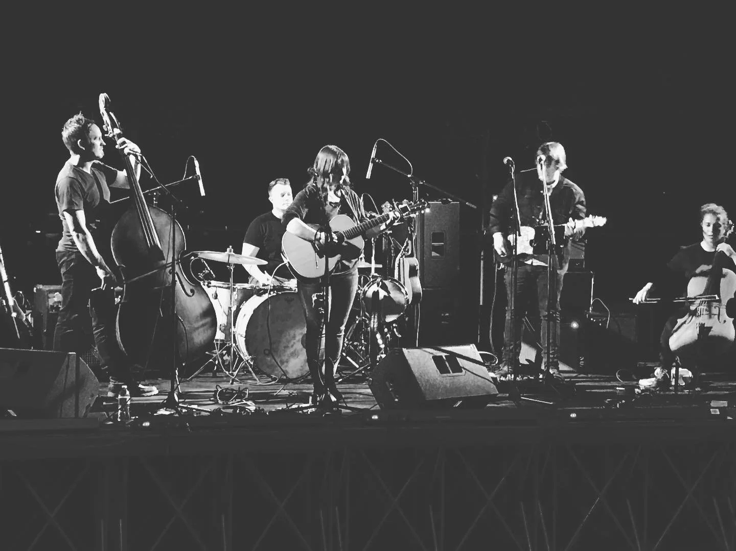 A black and white photo of a band performing on stage with five musicians: a bassist, a drummer, a guitarist singing into a microphone, a guitarist, and a cellist, all focused on their instruments.