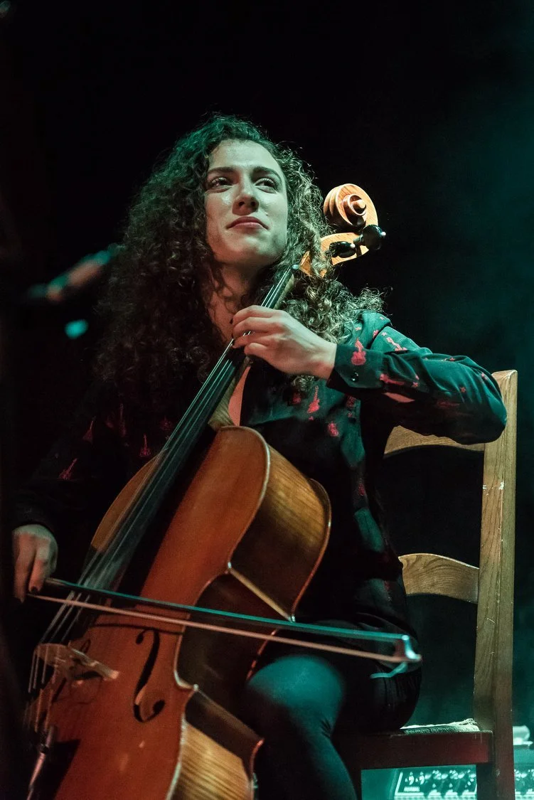 Natalie Mai Hall, a woman with curly hair playing a cello on stage, sitting on a wooden chair, with a focused expression.