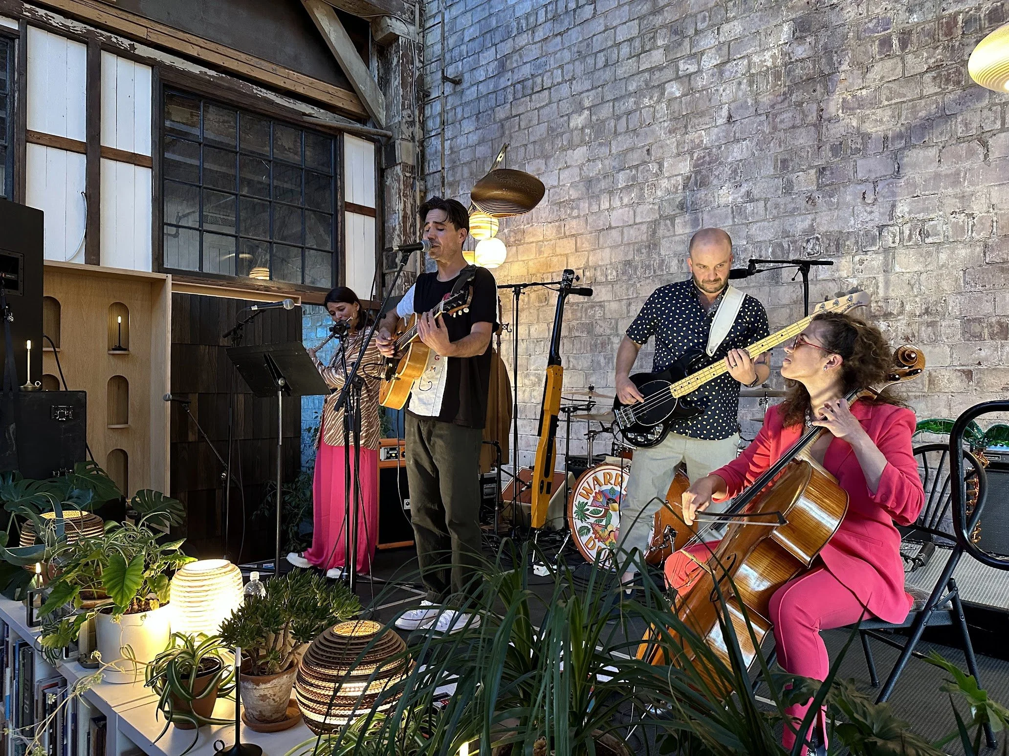 A four-member band performing indoors against a brick wall, with two female musicians and two male musicians playing guitar, bass, and cello, surrounded by plants and warm lighting.