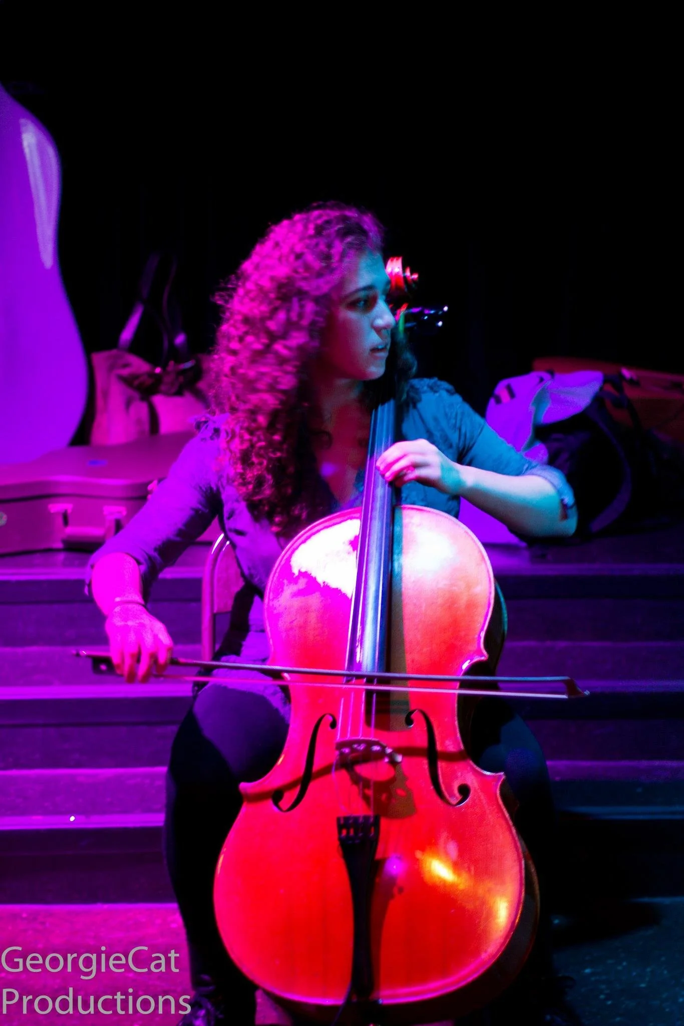 Natalie Mai Hall, a woman with curly hair playing a cello on stage, illuminated by purple and pink stage lights, with musical equipment and bags in the background.