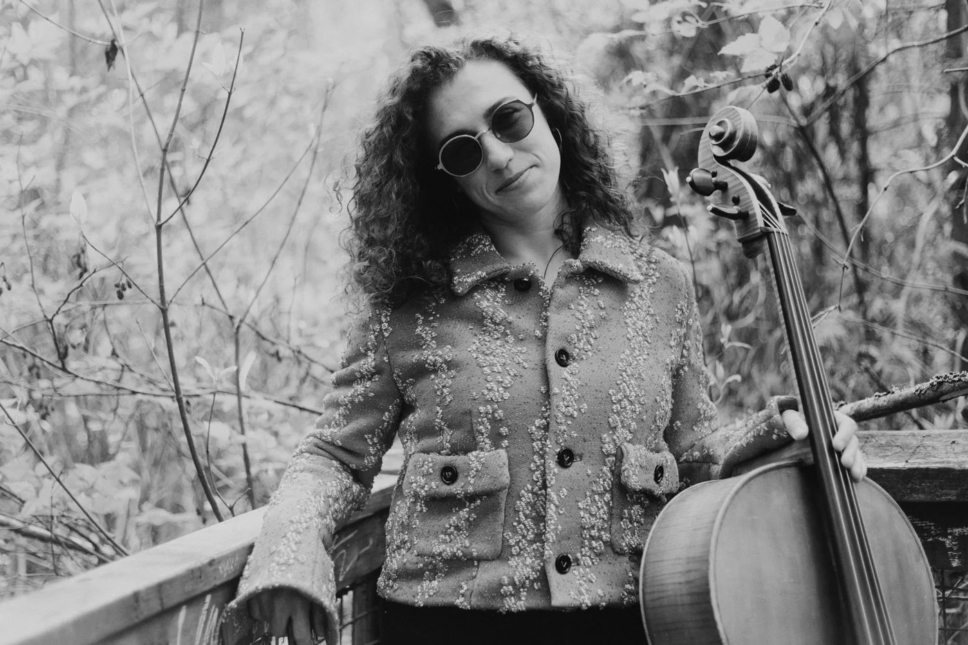 Natalie Mai Hall, a woman with curly hair wearing sunglasses and a textured jacket, is posing outdoors with a cello in a wooded area.