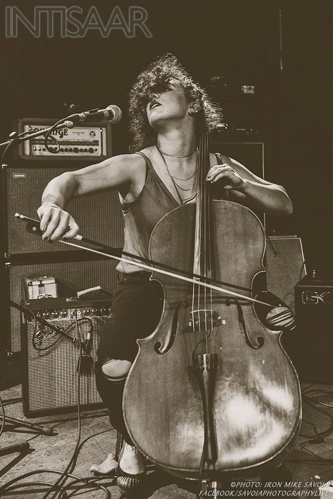 Natalie Mai Hall, a woman playing the cello on stage, has her eyes closed and her hair covering her face as the cello is run through an amplifier stack. 